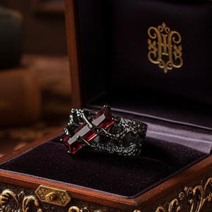 Può includere: Un anello nero con una croce di pietra preziosa rossa è esposto all'interno di un portagioie in legno ornato aperto. La scatola è marrone scuro con accenti color oro e un interno in velluto. La scatola è su una superficie a motivi floreali.