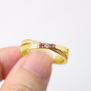 以下が含まれることがあります： 金色のリングで、クロスデザインです。リングには、小さなキラキラ光る白い石と、3つの宝石（ピンク2つとクリア1つ）が付いています。リングは人の手で持たれています。