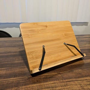 Puede incluir: Un atril de bambú con un acabado natural de vetas de madera. El soporte tiene una base rectangular y una parte trasera en ángulo, sostenida por brazos de metal negro. El soporte está diseñado para sujetar libros o tabletas.