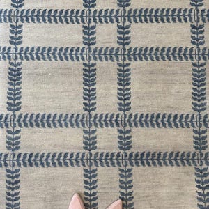 May include: Beige rug with a grid pattern of blue leaf-like designs. The rug has a textured appearance and is rectangular. The image also shows a pair of pink shoes.