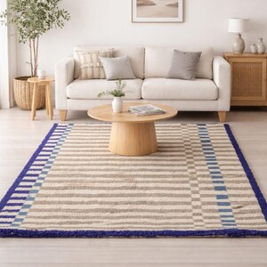 K&ouml;nnte beinhalten: Ein beige-blau gestreifter Teppich mit dunkelblauem Rand. Der Teppich hat horizontale Streifen in Beige und Grau, mit einem Abschnitt aus blauen und grauen Quadraten. Der Teppich befindet sich in einem Wohnzimmer.