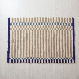 K&ouml;nnte beinhalten: Rechteckiger Teppich mit Streifenmuster in Beige und Grau, akzentuiert mit blauen Quadraten und einem blauen Rand. Der Teppich hat ein strukturiertes, weiches Aussehen, geeignet f&uuml;r ein Badezimmer oder einen Eingangsbereich.