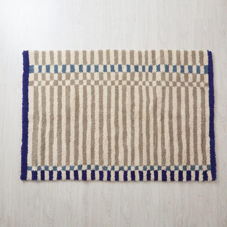 K&ouml;nnte beinhalten: Rechteckiger Teppich mit Streifenmuster in Beige und Grau, akzentuiert mit blauen Quadraten und einem blauen Rand. Der Teppich hat ein strukturiertes, weiches Aussehen, geeignet f&uuml;r ein Badezimmer oder einen Eingangsbereich.