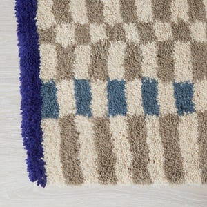K&ouml;nnte beinhalten: Nahaufnahme eines strukturierten Teppichs mit einem Schachbrettmuster in Beige, Grau und Blau. Der Teppich hat einen tiefblauen Rand. Das Design zeigt abwechselnde Quadrate und Streifen, wodurch ein geometrisches Muster entsteht.