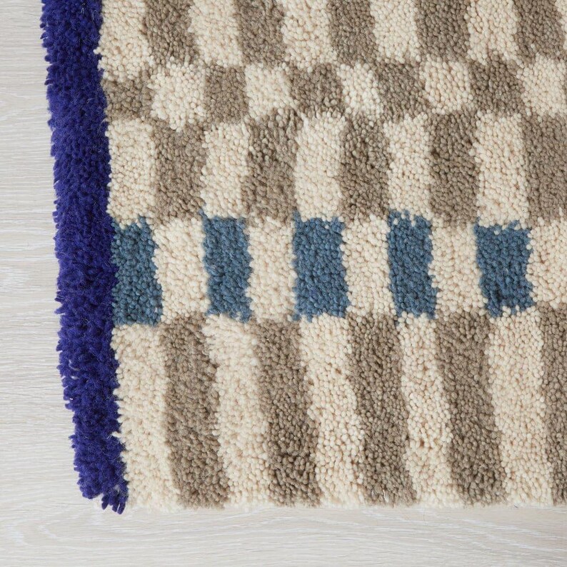K&ouml;nnte beinhalten: Nahaufnahme eines strukturierten Teppichs mit einem Schachbrettmuster in Beige, Grau und Blau. Der Teppich hat einen tiefblauen Rand. Das Design zeigt abwechselnde Quadrate und Streifen, wodurch ein geometrisches Muster entsteht.