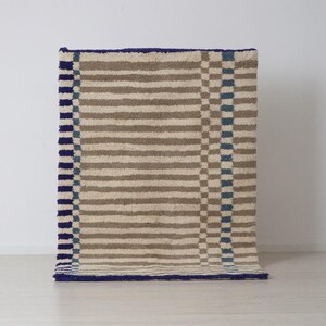 K&ouml;nnte beinhalten: Ein rechteckiger Teppich mit einem gestreiften Muster in Beige und Braun, mit blauem Rand und Akzenten. Der Teppich hat ein strukturiertes, weiches Aussehen, geeignet f&uuml;r ein Wohn- oder Schlafzimmer. Das Design umfasst vertikale Streifen und ein Schachbrettmuster.