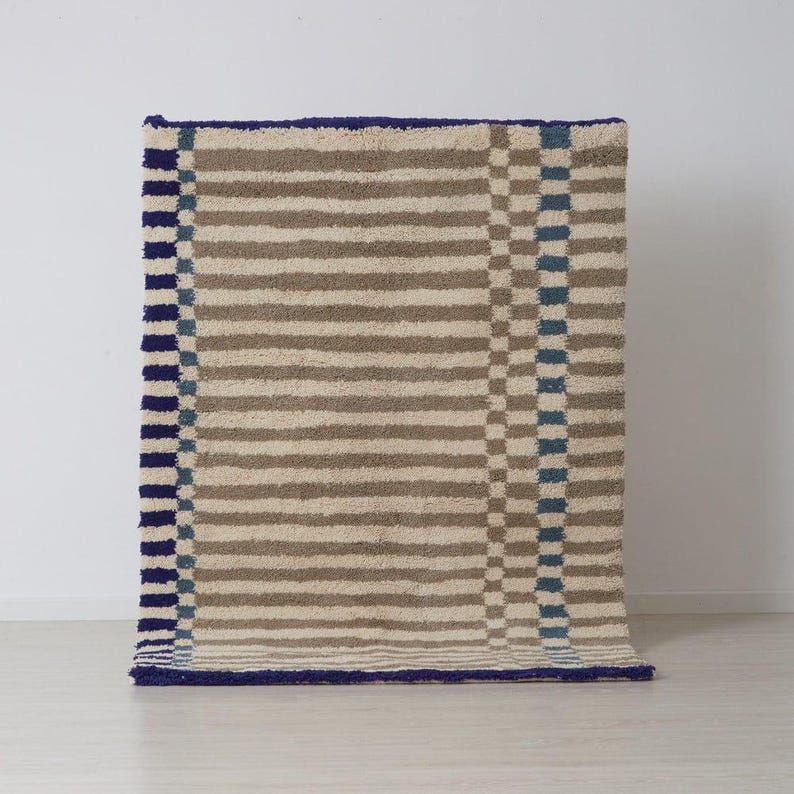 K&ouml;nnte beinhalten: Ein rechteckiger Teppich mit einem gestreiften Muster in Beige und Braun, mit blauem Rand und Akzenten. Der Teppich hat ein strukturiertes, weiches Aussehen, geeignet f&uuml;r ein Wohn- oder Schlafzimmer. Das Design umfasst vertikale Streifen und ein Schachbrettmuster.