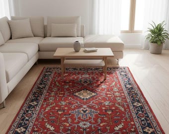 Könnte beinhalten: Ein rechteckiger roter Teppich mit einem aufwendigen, traditionellen Design. Der Teppich hat ein zentrales Medaillon und einen dunkelblauen Rand mit floralen Mustern. Er liegt in einem Wohnzimmer mit beigem Sofa und Holztisch.