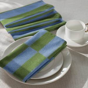 May include: A set of folded cloth napkins featuring a repeating pattern of wide horizontal stripes in light blue and olive green. The napkins are displayed alongside a white ceramic teacup and saucer set resting on a stack of white dinner plates.
