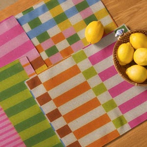 Op de afbeelding: Een verzameling kleurrijke placemats met geometrische patronen. De placemats hebben strepen en rechthoekige blokken in roze, groen, oranje en blauw. Een mandje met citroenen is ook zichtbaar.