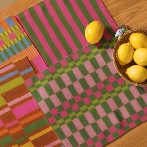 May include: A collection of colorful placemats with geometric patterns in pink, green, orange, and blue. A woven basket holds several yellow lemons. The placemats are arranged on a wooden surface, creating a vibrant and inviting scene.
