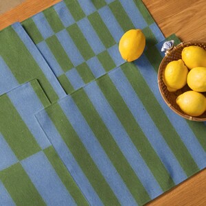 May include: Two blue and green striped placemats with a checkerboard pattern. A small woven basket filled with yellow lemons sits on the right side of the image. One lemon is placed on the placemat.