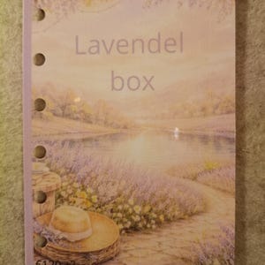 Op de afbeelding: Een kaart met de tekst "Lavendel box". De afbeelding toont een landschap met een pad, een meer en een hoed. Het kleurenpalet is zacht, met tinten paars, beige en groen.