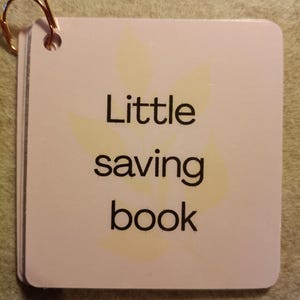 Könnte beinhalten: Ein kleiner Stapel weißer Karten mit abgerundeten Ecken, zusammengehalten von einer goldfarbenen Büroklammer. Die Karten zeigen die Worte "Little saving book" in Schwarz, mit einem blassgelben Blattmuster.