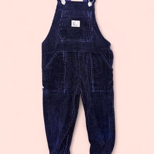 May include: Navy blue corduroy overalls with adjustable straps and metal hardware. The overalls have a front pocket with a small white label and two side pockets. The cuffs are black. The overall design is a classic style.