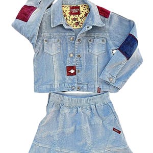 Könnte beinhalten: Hellblaues Cord-Jacke- und Rock-Set. Die Jacke hat bordeauxfarbene Patches auf den Schultern und marinefarbene Patches auf den Ärmeln. Jacke und Rock tragen die Marke "GUESS". Die Jacke hat ein floral gemustertes Futter.