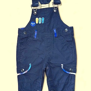May include: Navy blue baby overalls with adjustable straps and button closures. The overalls feature a patch with the text "Zippy" and colorful surfboard designs. The pockets have a blue and white plaid trim. The overall is made of a soft material.