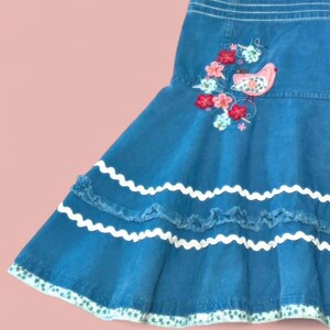 Peut inclure: Jupe en jean bleu avec un ourlet à volants et une bordure blanche en zigzag. La jupe présente un motif brodé d'oiseaux et de fleurs roses, rouges et bleues. L'ourlet est orné d'un motif floral vert et blanc.