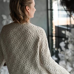 May include: Cream-colored knit sweater with a detailed, repeating pattern. The sweater has a crew neck and long sleeves. The intricate design covers the entire garment, creating a textured appearance. The model is wearing a brown skirt.