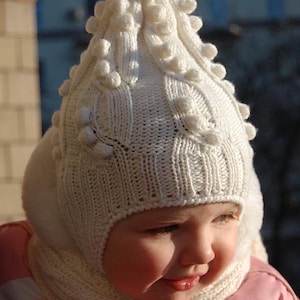 Könnte beinhalten: Ein weißer Strick-Winterhut mit passendem Schal. Der Hut hat ein strukturiertes Design mit Bommeln und Ohrenklappen. Das Kind trägt eine rosa Jacke. Das Set ist aus weichem, warmem Material gefertigt.