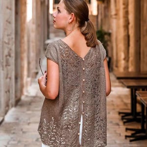 May include: A brown, open-back knit top with a V-neck and cap sleeves. The back features a button-up closure and a delicate, openwork design. The top is worn over white pants.