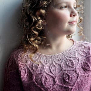 May include: A pink knitted jumper with a leaf design across the yoke and chest. The jumper has a round neckline and long sleeves. The model is wearing a beaded headband and has curly hair. The jumper is made of wool.
