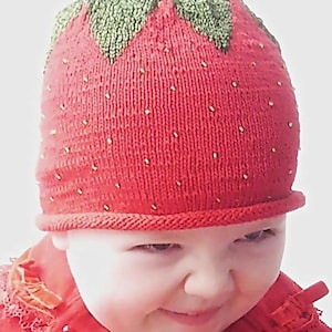 Könnte beinhalten: Eine gestrickte rote Erdbeermütze mit einem grünen, blattförmigen Oberteil und kleinen goldfarbenen Verzierungen. Die Mütze wird von einem Baby getragen. Die Mütze hat einen gerollten Rand. Das Baby trägt eine rote Jacke. Die Website www.knitting.com.ua ist unten sichtbar.