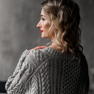 Pode incluir: Uma mulher com cabelo loiro e ondulado usando um suéter cinza de malha de cabo. O suéter tem um padrão texturizado com detalhes grossos e trançados. A mulher é vista de costas e de lado, com batom vermelho.