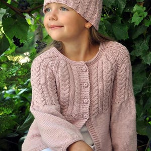 Puede incluir: Conjunto de gorro y cárdigan de punto rosa claro. El gorro y el cárdigan presentan un diseño de punto trenzado. El cárdigan tiene una botonadura frontal. El conjunto está hecho de un material suave y cálido.