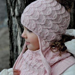 May include: A light pink knitted hat and scarf set with a cable knit pattern. The hat has ear flaps and a brown fur pom-pom on top. The scarf is a matching color and design. The set is designed for cold weather.