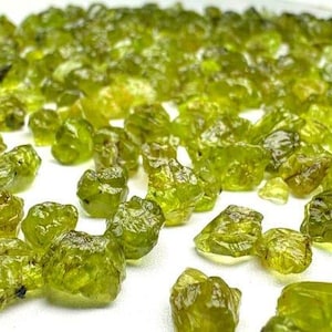 Op de afbeelding: Een verzameling kleine, ruwe, doorschijnende olijfgroene stenen met onregelmatige, grillige vormen en gefacetteerde oppervlakken. De stenen variëren in grootte en vertonen een levendige, halfdoorzichtige kwaliteit, verspreid over een effen, helderwit oppervlak.