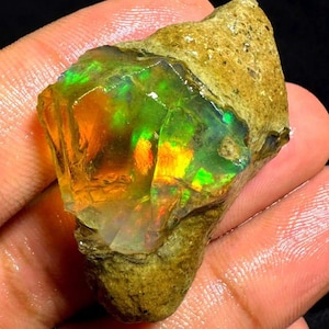 Op de afbeelding: Een ruwe, ongepolijste opaal edelsteen met een ruwe, getextureerde beige en bruine buitenmatrix. Het blootgestelde kristallijne gedeelte vertoont levendige flitsen van iriserend groen, oranje en geel licht, wat een kleurenspel-effect creëert.
