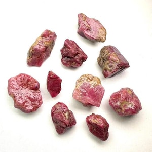 May include: A collection of ten raw, unpolished ruby gemstones in varying sizes and irregular shapes. Each stone displays a deep magenta to dark pink hue with natural surface textures, including patches of earthy brown and tan mineral inclusions.