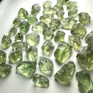 Peut inclure: Une collection de nombreuses pierres de verre brutes et translucides, de couleur vert clair, aux formes irrégulières, dentelées et aux bords tranchants. Chaque pièce présente une surface facettée unique qui capte la lumière.