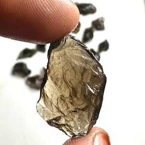 May include: A close-up of a rough, translucent, brown gemstone held between two fingers. Several other similar stones are scattered in the background. The gemstone has a textured surface and irregular edges, catching the light.