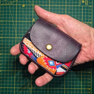 May include: A small, purple leather wallet with a patterned fabric front. The fabric features a colourful geometric design in red, orange, blue, and white. The wallet has a snap closure and is held in a hand.