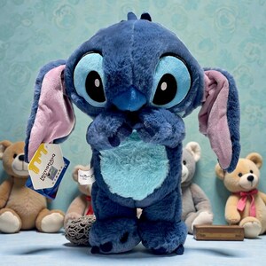 May include: A plush Stitch doll in a standing pose, featuring large black and blue eyes, blue fur, and pink-lined ears. The doll has a light blue belly and is holding its hands together. A tag is attached to the doll.