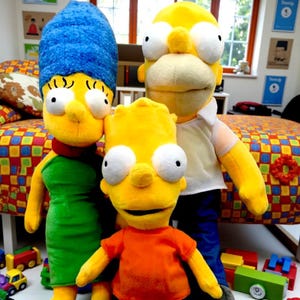 May include: Three plush dolls representing the Simpson family: Marge, Homer, and Bart. Marge is in a green dress with blue hair. Homer wears a white shirt and has yellow skin. Bart is in an orange shirt. The dolls are in a child's room.