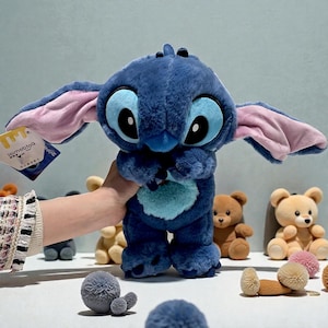 May include: A plush blue Stitch doll with large eyes, ears with pink inner lining, and a light blue heart-shaped patch on its chest. The doll is held in front of a white surface with other plush toys.