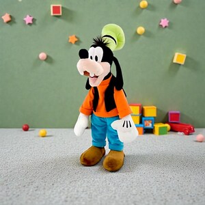 May include: A plush toy of Goofy, a Disney cartoon character, stands on a gray surface. Goofy wears an orange shirt, blue pants, brown shoes, and a green hat. His black ears and black vest are visible. White gloves complete the outfit.