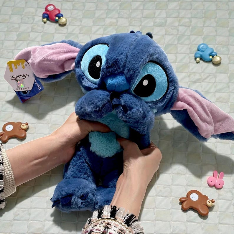 May include: A plush blue Stitch character with large black eyes and pink-lined ears. The plush toy is held by hands and has a tag attached. Small decorative figures are scattered around the plush toy.