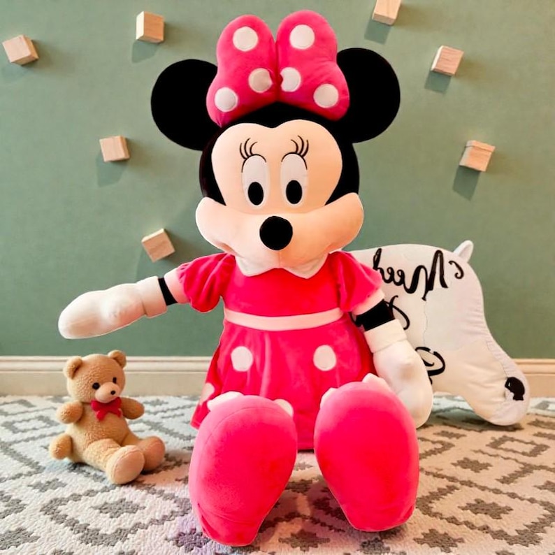 May include: A large Minnie Mouse plush doll wearing a pink dress with white polka dots and a matching bow. A small teddy bear sits nearby. The doll has black ears and a white face.