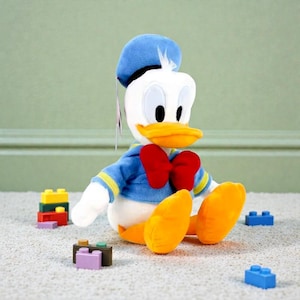 May include: A plush Donald Duck toy, featuring a blue sailor shirt, hat, and a red bow tie. The toy has a white face, yellow beak and feet. Several colorful toy blocks are scattered around the plush toy.