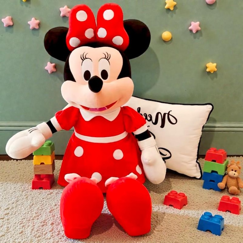 May include: A large Minnie Mouse plush toy, wearing a red dress with white polka dots and a matching bow. The plush has black ears and a white collar. The toy is sitting on a light-colored carpet, surrounded by building blocks and a pillow.