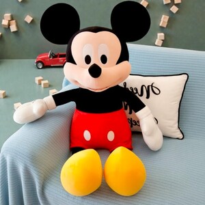 May include: A plush Mickey Mouse doll with a black torso and arms, red shorts, and yellow feet. The doll has large black ears, white gloves, and a smiling face. A red toy car and a white pillow are in the background.