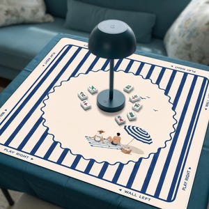 May include: A square mahjong mat with a blue and white striped border and a central cream-colored circle featuring a beach scene with two figures and an umbrella. The mat includes the text PLAY RIGHT and WALL LEFT. A dark blue lamp sits in the center.