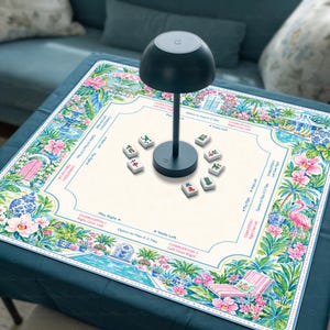 May include: A square mahjong mat featuring a colorful botanical border with pink flowers, green palm leaves, and a flamingo, surrounding a cream-colored center with game instructions and text. A dark gray mushroom-shaped lamp sits in the middle.