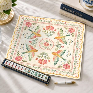 May include: A square mahjong mat featuring a cream background with a floral and bird pattern in shades of pink, green, and orange. A dark blue tile rack holds white mahjong tiles with red and green characters and symbols.