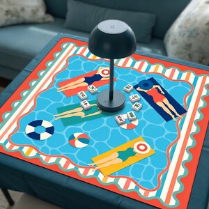 May include: A square table mat featuring a blue swimming pool pattern with figures on towels, beach balls, and scattered mahjong tiles. A dark gray mushroom-shaped lamp with a power button sits in the center of the mat on a dark blue surface.