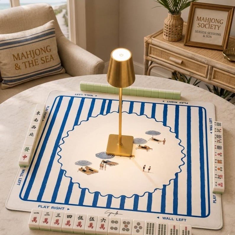 Puede incluir: Un tapete de mahjong cuadrado con un borde de rayas blancas y azules y una ilustraci&oacute;n circular central de escenas de playa con sombrillas y tumbonas. Una l&aacute;mpara de metal dorado se encuentra en el centro, rodeada de fichas de mahjong.
